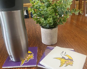 May include: A stainless steel travel mug rests on a purple and white tile coaster featuring the Minnesota Vikings logo. A small potted plant with green leaves sits nearby. The coasters are square.