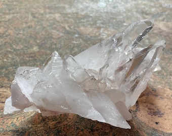 May include: A cluster of clear quartz crystals with a natural, raw appearance. The crystals are arranged in a cluster, with some pointing upwards and others pointing downwards. The crystals are translucent and have a slight pink hue.