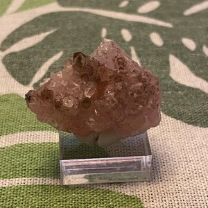 May include: A cluster of pink amethyst crystals with a brown hue, displayed on a clear acrylic stand.