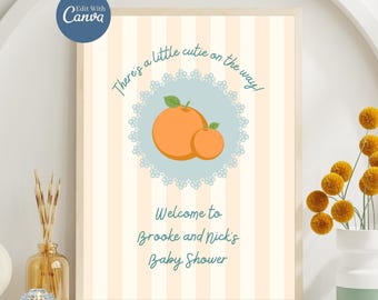 May include: A framed baby shower sign with a light orange and white striped background. The sign features the text "There's a little cutie on the way!" and "Welcome to Brooke and Nick's Baby Shower." An illustration of two oranges is in the center.