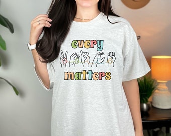 Op de afbeelding: Een grijs T-shirt met de tekst "every voice matters" in kleurrijke letters. De tekst wordt vergezeld door een afbeelding van vijf handen in verschillende gebarentaalposes, verbonden door een touwtje.