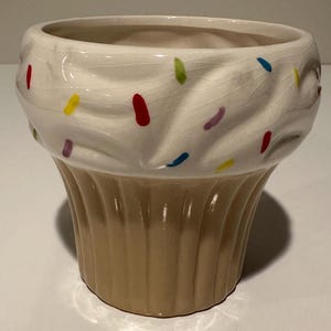 Pode incluir: Um recipiente de cerâmica em forma de cupcake com um topo creme e uma base bege. O topo é decorado para se assemelhar a cobertura, com granulado colorido. O recipiente foi projetado para se parecer com um cupcake.