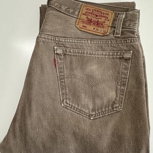 May include: Folded brown Levi's 501 jeans. The jeans have a classic five-pocket design, a red Levi's tag on the back pocket, and a brown leather patch with the Levi Strauss & Co. logo. The waist size is W34 and the length is L32.