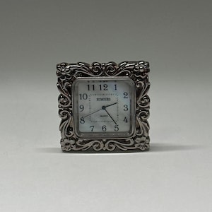 May include: A small, square, silver-toned clock with an ornate floral and scrollwork frame. The clock face is white with black numerals and hands, and the word "RUMOURS" is printed on the face. The clock is a decorative item.