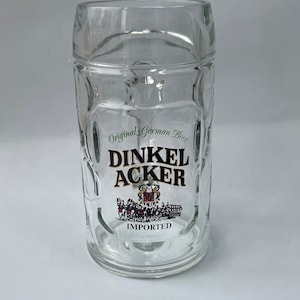 May include: Clear glass beer stein with a rectangular body and a handle. The stein features the words "DINKEL ACKER" in a black and gold font, with "Original German Beer" above and "IMPORTED" below. A colorful emblem is also displayed.