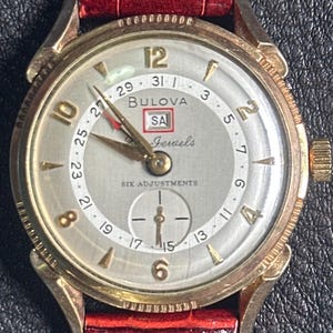 May include: A vintage Bulova wristwatch with a gold-tone case and a light-colored face. The watch features gold hands, hour markers, and a red leather band. The face displays the Bulova logo, the text "SA Jewels", and "SIX ADJUSTMENTS".