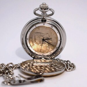 May include: Silver pocket watch with a chain, open to reveal a gold-toned face with Roman numerals. The watch has a decorative top and a detailed back. The chain is silver and attached to the watch.