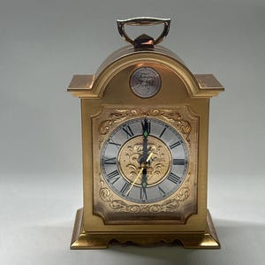 May include: A gold-toned, rectangular clock with a handle on top. The clock face has Roman numerals and ornate gold detailing. The words "Tempus Fugit" are displayed above the clock face. The clock is a decorative item.