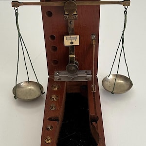 May include: A vintage wooden balance scale with a hinged lid. The scale features a brass arm, two metal pans suspended by green cords, and a small measurement gauge. The wooden box is a dark brown color.