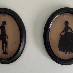May include: Two oval framed silhouettes. One depicts a man in a coat and holding a cane, the other a woman in a gown. The black silhouettes are set against a tan background, framed in black.