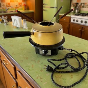 May include: A yellow electric fondue pot with a black handle, resting on a black circular heating base with a temperature control dial and a black power cord. Three metal fondue forks with dark brown handles are inserted into the pot.