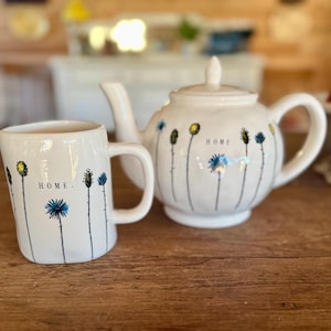 May include: White ceramic teapot and mug set. The teapot has a lid and spout, and the mug has a handle. Both pieces are decorated with black stems and yellow and blue flower designs. The word "HOME" is printed on both items.
