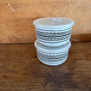 May include: Two stacked white ceramic bowls with clear plastic lids. Each bowl features a black geometric pattern consisting of horizontal lines, dashes, and zig-zag shapes encircling the exterior. The bowls are round with a smooth finish.