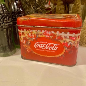 May include: A vintage red Coca-Cola tin with a floral and picket fence design. The tin features the Coca-Cola logo and the words "Delicious and Refreshing". The tin is rectangular and has a slightly worn appearance.