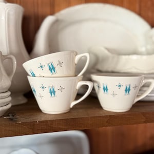 May include: A stack of three white teacups with a turquoise and black geometric pattern. The cups are on a wooden shelf with other white dishware in the background. The cups have a curved handle.