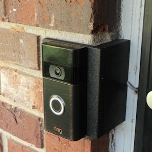 May include: A bronze Ring video doorbell fixed to a red brick wall. The doorbell features a camera lens, a white-ringed button, and the brand name "ring". A black mounting bracket is attached to the side, next to a door frame.