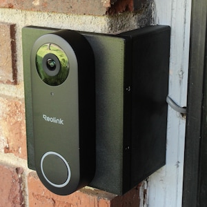 May include: A black Reolink video doorbell mounted on a red brick wall. The doorbell has a camera lens, a circular button, and the Reolink logo. The doorbell is installed on a black mounting bracket.
