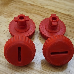 Puede incluir: Cuatro perillas de plástico rojo con un borde estriado y un poste central. Dos perillas tienen una ranura y dos tienen una parte superior en forma de cruz. Las perillas están dispuestas sobre una superficie de madera.