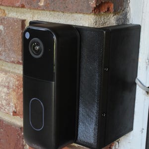 May include: A black smart doorbell camera mounted on a red brick wall. The camera has a lens, a blue light, and a button. The doorbell is attached to a black mounting bracket.