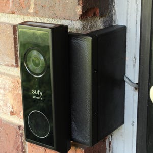 May include: A black eufy Security doorbell camera mounted on a red brick wall. The camera has a circular lens and a rectangular design. The doorbell is installed next to a white door frame.