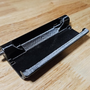 Könnte beinhalten: Eine schwarze, 3D-gedruckte Kunststoff-Scharnierkomponente mit einer rechteckigen Basis, einem erhöhten Mittelkanal und einer metallischen Gewindeschraube, die an einer Seite herausragt, konzipiert für mechanische Montage und Stützzwecke.
