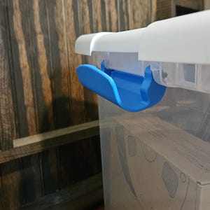 Puede incluir: Un contenedor de almacenamiento de plástico transparente con una tapa blanca y un mecanismo de cierre de color azul brillante. El contenedor es rectangular con un cuerpo liso y transparente, y el pestillo azul está fijado al lateral.