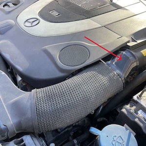 May include: An engine compartment with a gray engine cover and a black air intake hose. The hose has a textured, corrugated design. The engine cover has a Mercedes-Benz logo.