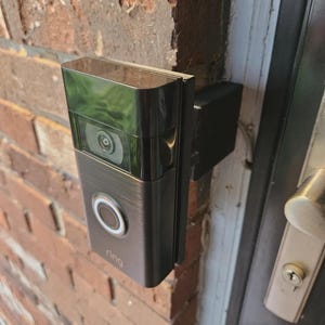 Puede incluir: Un timbre de video Ring negro y plateado montado en una pared de ladrillos rojos. El timbre tiene una lente de cámara y un botón circular con un anillo blanco brillante. La palabra "ring" está impresa debajo.