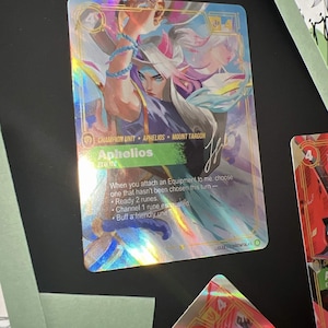 May include: A rectangular holographic trading card featuring the character Aphelios from Mount Targon. The card displays the text Champion Unit, Aphelios, and game instructions including Ready 2 runes, Channel 1 rune exhausted, and Buff a friendly unit.