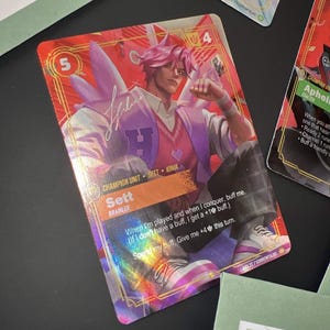 May include: A holographic trading card featuring a character named Sett with pink hair, wearing a letterman jacket over a pink sweater. The card displays the text 'Champion Unit', 'Sett', 'Brawler', and game mechanics text in a red and orange frame.