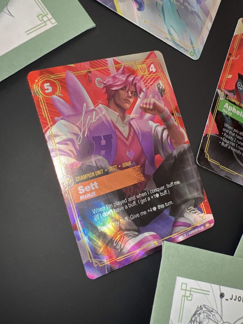 May include: A holographic trading card featuring a character named Sett with pink hair, wearing a letterman jacket over a pink sweater. The card displays the text 'Champion Unit', 'Sett', 'Brawler', and game mechanics text in a red and orange frame.