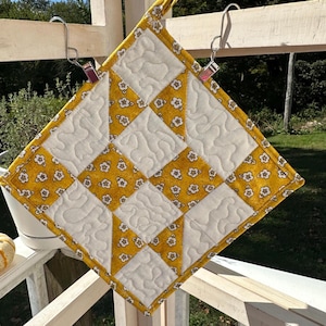 May include: A square, quilted potholder with a yellow and white floral pattern. The potholder is made of fabric and has a diamond pattern. It is hanging from a metal hook.