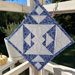 May include: A square, quilted pot holder with a blue and white geometric pattern. The pot holder is made of fabric and has a loop for hanging. The design features white floral shapes and blue speckled triangles.