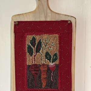 May include: A wooden cutting board with a red fabric panel featuring a needlepoint design of green leafy vegetables and orange and red carrots.