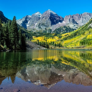 May include: A scenic view of a mountain lake with a reflection of the surrounding mountains and trees. The lake is surrounded by lush green trees and the mountains are covered in snow. The water is calm and clear, reflecting the colors of the sky and the trees.