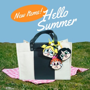 May include: A white canvas tote bag with a black leather panel and a zipped pocket. The bag has three colourful crocheted character charms attached to the zipper pull. The text "New Items! Hello Summer" is in the background.