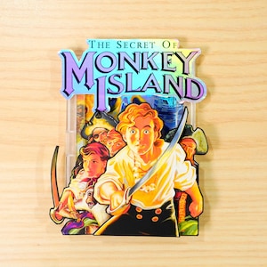 May include: A vibrant, holographic sticker with the words "The Secret of Monkey Island." The image shows a young man with a sword, surrounded by other characters, and a pirate ship. The sticker is placed on a wooden surface.