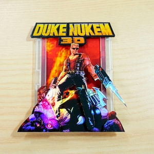 Può includere: Un espositore per cartucce di gioco Duke Nukem 3D stampato in 3D. L'opera d'arte presenta il logo di Duke Nukem e un personaggio muscoloso che tiene in mano una pistola. Lo sfondo è di un rosso vibrante e il design generale è un omaggio al videogioco classico.