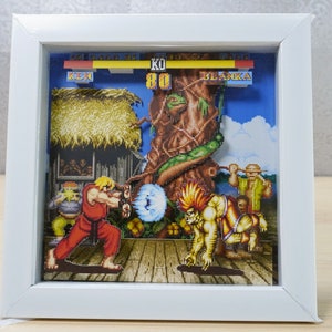 Puede incluir: Un diorama 3D de una escena de videojuego con dos personajes, Ken y Blanka, de la serie de videojuegos Street Fighter. El diorama está ambientado en un entorno de jungla con un gran árbol y una cabaña de paja. Los personajes están en posición de combate, con Ken en un gi rojo y Blanka en un atuendo verde y amarillo. El diorama está enmarcado en un marco blanco.