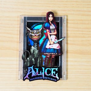 Può includere: Una custodia di plastica trasparente con una grafica del videogioco "Alice: Madness Returns". L'immagine mostra Alice in un vestito blu che tiene un coltello, insieme allo Stregatto. Il titolo del gioco è visualizzato in un font stilizzato.