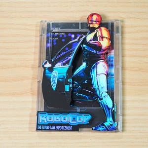 Puede incluir: Figura de acción de Robocop en una caja de plástico transparente. La figura lleva un uniforme de policía futurista, de pie junto a un coche de policía. El texto "ROBOCOP" y "THE FUTURE LAW ENFORCEMENT" son visibles.