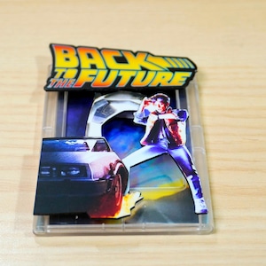 Puede incluir: Un objeto de colección con el tema de Regreso al Futuro, con el logotipo de la película y una ilustración de personaje. La imagen incluye un coche DeLorean y un personaje en una pose dinámica, sobre un fondo futurista. El diseño está encerrado en una funda de plástico transparente.