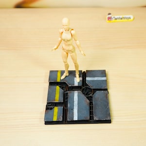 Op de afbeelding: Een beige beweegbaar actiefiguur dat op een vierkante, donkergrijze displaybasis staat. De basis heeft een verweerd oppervlak in industriële stijl met geschilderde gele en witte lijnen, die doen denken aan een straat of platform.