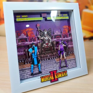 Puede incluir: Un diorama 3D enmarcado en blanco que representa una escena del videojuego Mortal Kombat II. El diorama presenta dos personajes, Sub-Zero y Mileena, luchando en un fondo morado y naranja. El diorama también incluye el título del juego, "Mortal Kombat II", en una fuente pixelada.