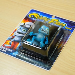 Könnte beinhalten: Eine blaue Crazy Frog-Actionfigur in einer transparenten Kunststoffverpackung. Die Verpackung zeigt das Crazy Frog-Logo und den Text "Axel F". Der Hintergrund zeigt ein Cartoon-Bild des Frosches. Die Verpackung liegt auf einer Holzoberfläche.
