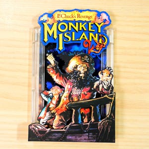 May include: A colorful, layered artwork featuring the text "Monkey Island 2" and a pirate theme. The artwork depicts a pirate captain with a skull-and-crossbones hat, a ship, and other characters. The background is blue.