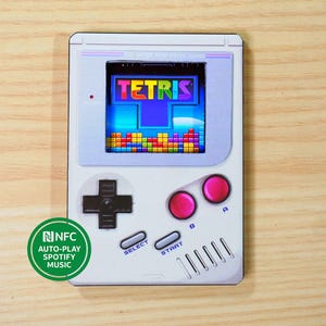May include: A white handheld gaming device featuring the Tetris game on the screen. The device has a directional pad, pink buttons, and the words "SELECT" and "START". A green NFC sticker indicates auto-play Spotify music.