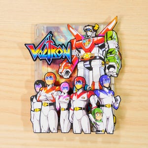 Op de afbeelding: Een kleurrijk Voltron-kunstwerk met de Voltron-robot en zijn team van piloten. Het kunstwerk bevat het Voltron-logo in een regenboogverloop, met de robot in rood, wit en groen. De piloten zijn in verschillende kleuren.