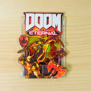 May include: A clear plastic case featuring the "DOOM Eternal" video game logo and artwork. The artwork depicts a muscular figure in orange armor wielding a weapon, with other characters and fiery elements.