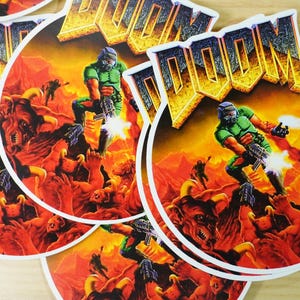 May include: Circular stickers featuring the word "DOOM" in a gold and white font. The stickers depict a green-armored character firing a weapon in a hellish landscape with demons. The stickers are round with a white border.